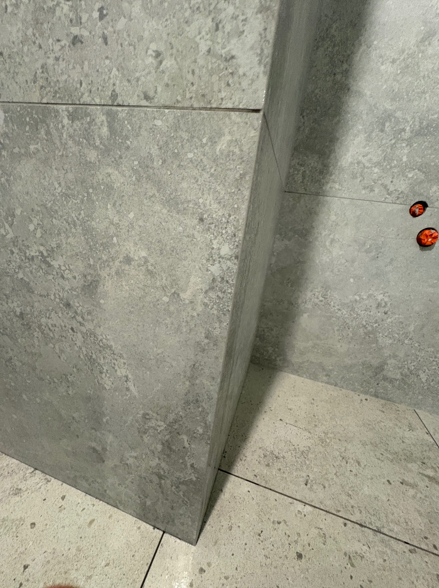 Szara, matowa ściana i podłoga wyłożona płytkami imitującymi beton. Widoczne gniazdka elektryczne. Minimalistyczne wykończenie wnętrza, surowy styl.