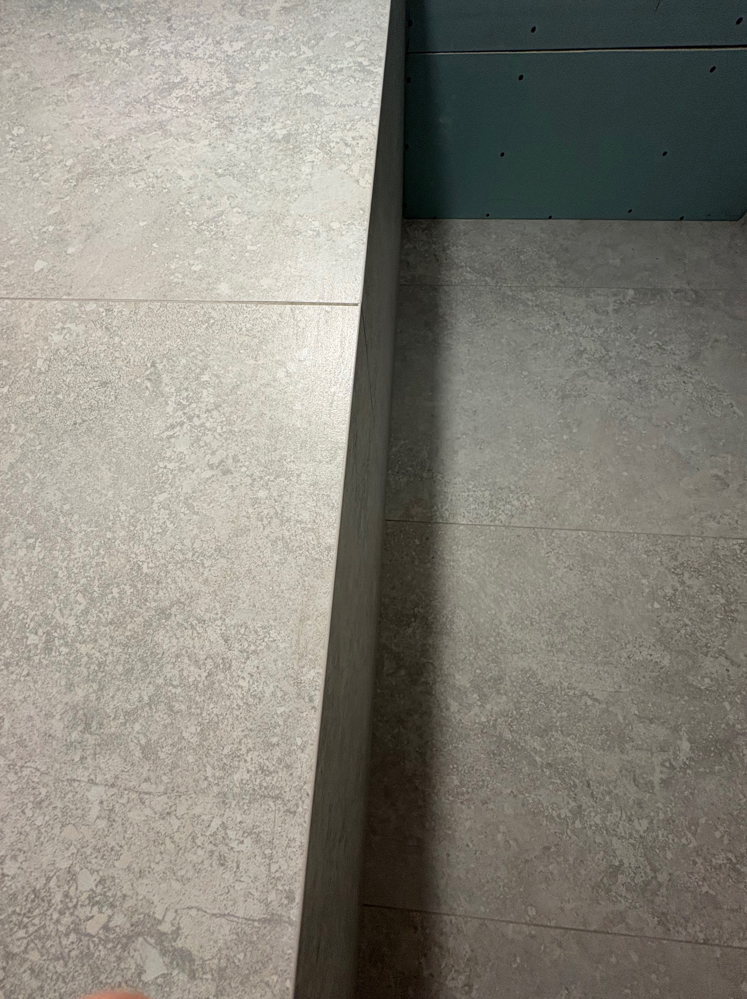 Szare płytki podłogowe z fugami i fragment ściany z płyty gipsowej. Widok z góry, bliskie ujęcie, neutralne oświetlenie.