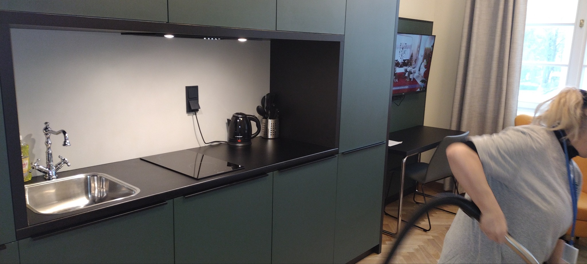 Kobieta odkurza podłogę w nowoczesnej kuchni z zielonymi szafkami i czarnym blatem. Widoczny zlew, płyta indukcyjna i czajnik elektryczny. W tle stół i krzesła.