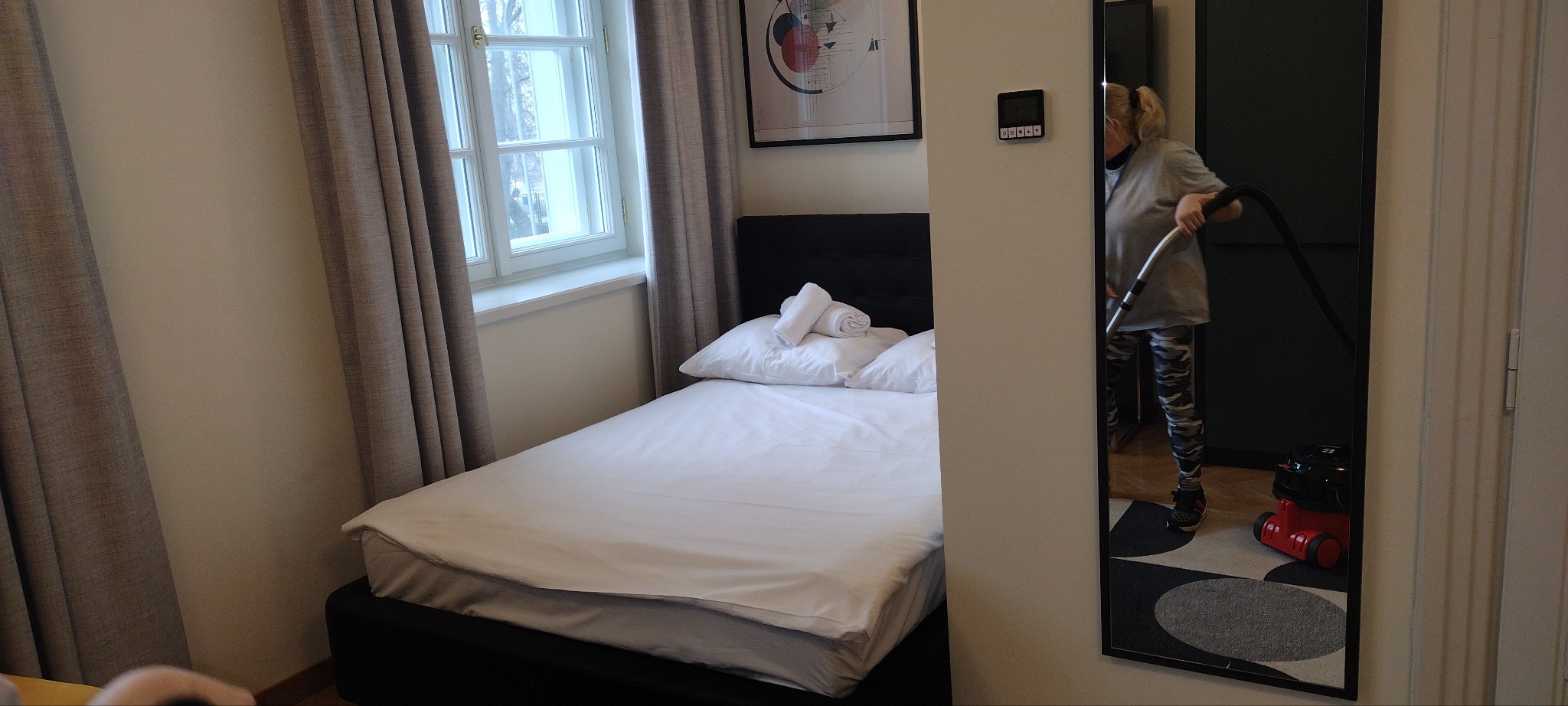 Pokój hotelowy z łóżkiem i oknem. W lustrze odbija się osoba odkurzająca podłogę czerwonym odkurzaczem. Widoczny obraz na ścianie i zasłony w oknie.