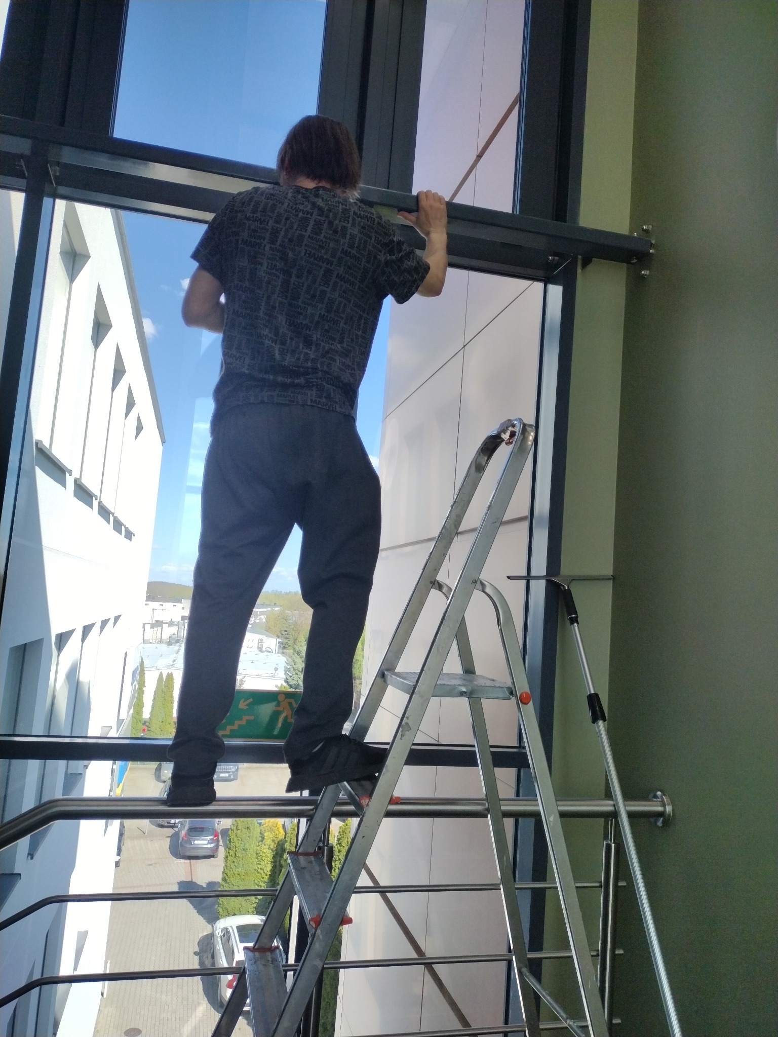 Mężczyzna na drabinie myje okna w biurowcu. Widoczne narzędzia do mycia okien i fragment budynku z zewnątrz. Słoneczny dzień.