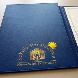 K.B. sp. z o.o. - Granatowa teczka z tłoczonym logo szkoły podstawowej i miejscem na dyplom, leżąca na jasnym tle. Widoczna faktura materiału i złote detale.
