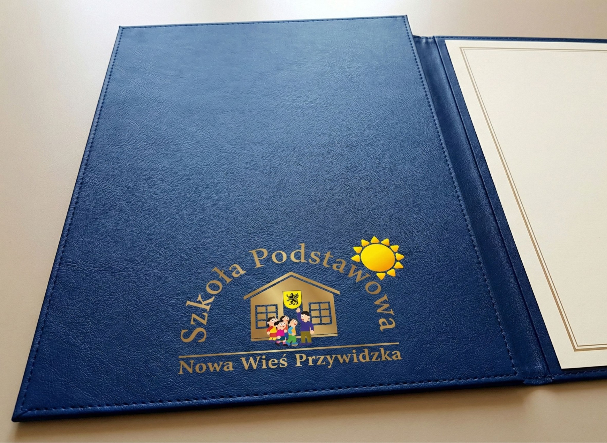 Granatowa teczka z tłoczonym logo szkoły podstawowej i miejscem na dyplom, leżąca na jasnym tle. Widoczna faktura materiału i złote detale.