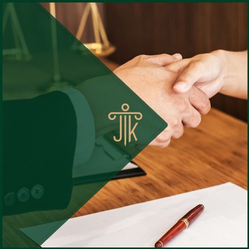 Uścisk dłoni nad dokumentami na drewnianym biurku, w tle waga sprawiedliwości. Częściowo zasłonięte logo kancelarii w lewym górnym rogu.