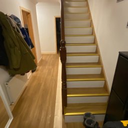 Montaż paneli winylowych na stopniach z białą podstopnicą i złotą listwą wykańczającą.