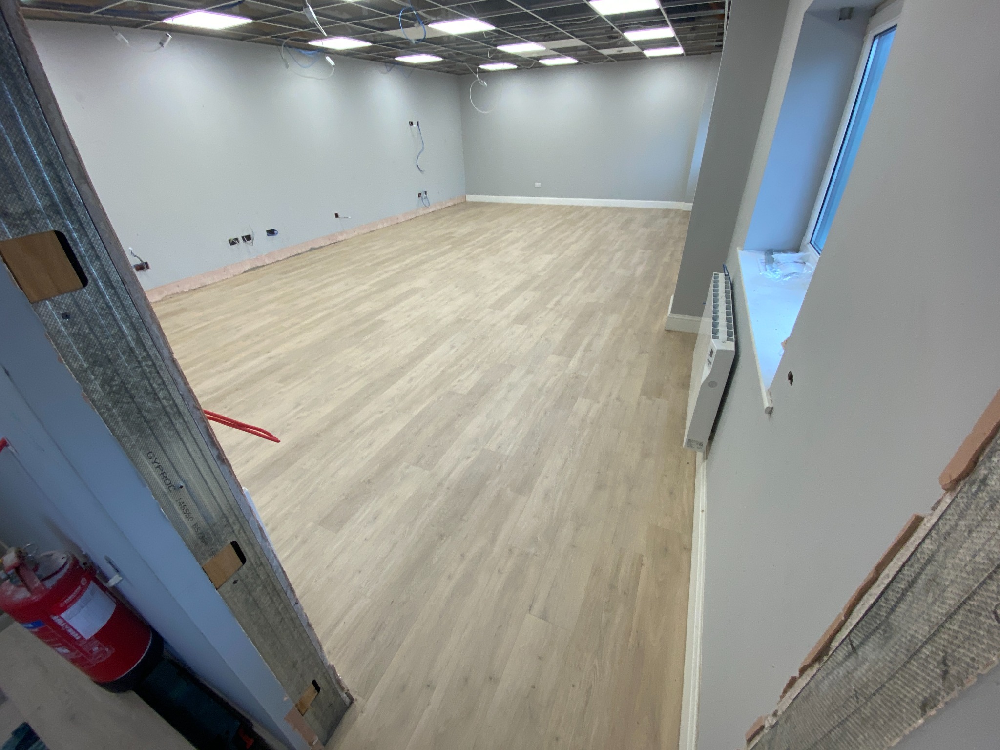 Wnętrze po remoncie: jasna podłoga, szare ściany, sufit z instalacją. Widoczny fragment ściany z płyt karton-gips i gaśnica. Okno z grzejnikiem.