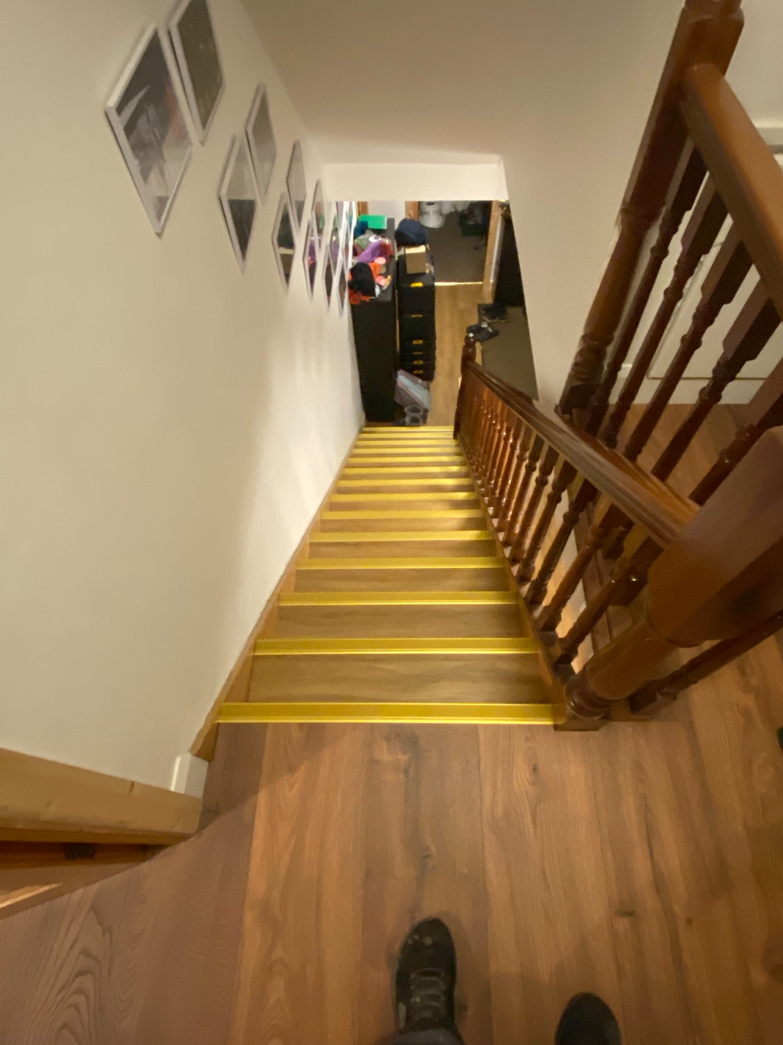 Widok z góry na drewniane schody z żółtymi paskami antypoślizgowymi, prowadzące w dół. Na ścianie galeria zdjęć w białych ramkach. Widoczne fragmenty podłogi i balustrady.