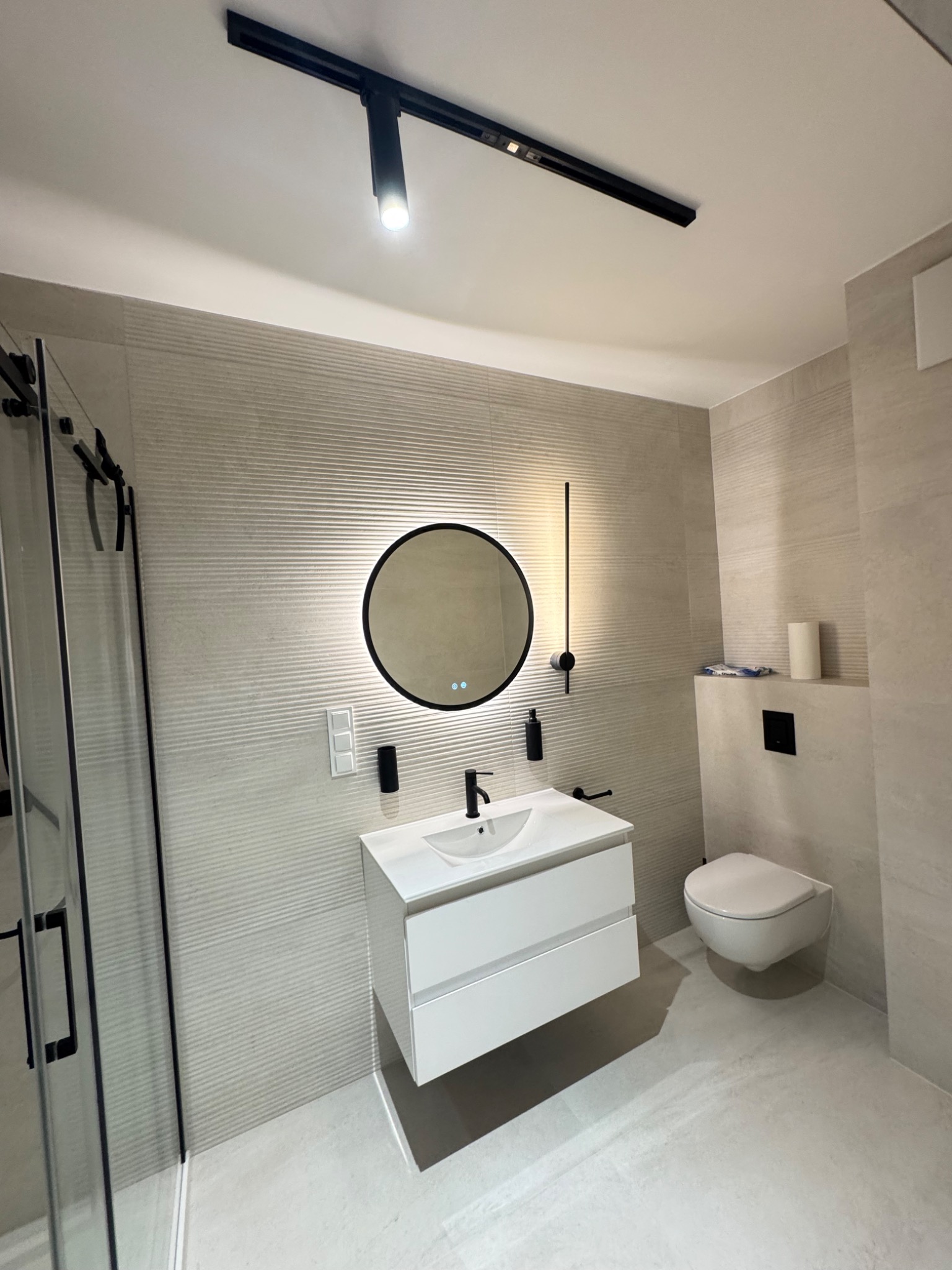 Nowoczesna łazienka w beżach: kabina prysznicowa z czarnymi ramami, okrągłe lustro z podświetleniem, biała szafka z umywalką i podwieszana toaleta. Minimalistyczne oświetlenie.