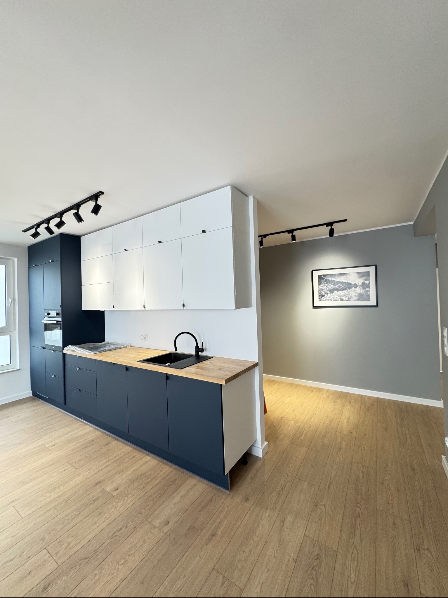 Nowoczesna kuchnia z granatowymi szafkami dolnymi i białymi górnymi, drewniany blat, czarna armatura, oświetlenie szynowe i parkiet. Minimalistyczny design.