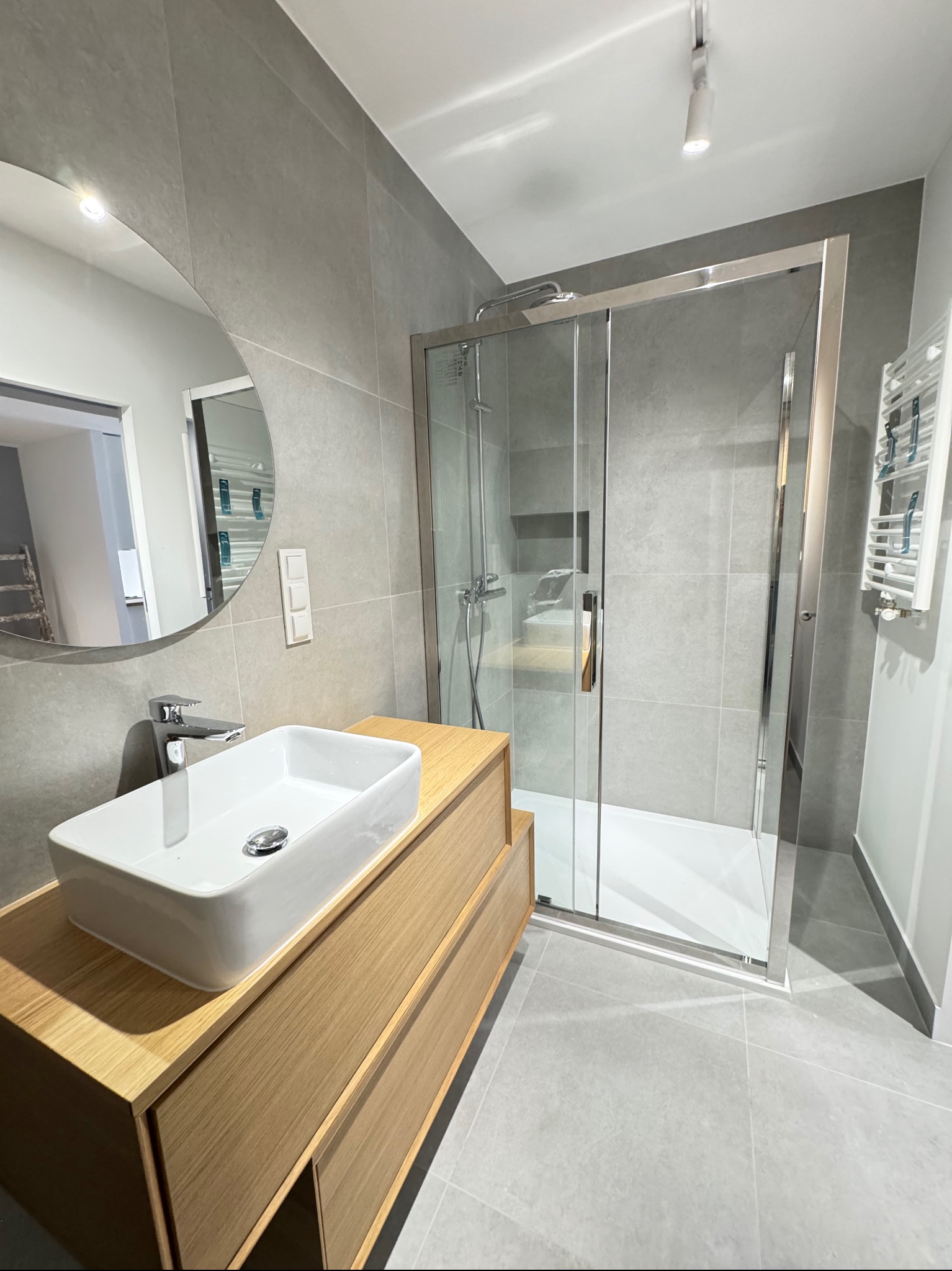 Nowoczesna łazienka w szarościach z drewnianą szafką pod umywalką i kabiną prysznicową ze srebrnymi profilami. Minimalistyczny design, okrągłe lustro i subtelne oświetlenie.