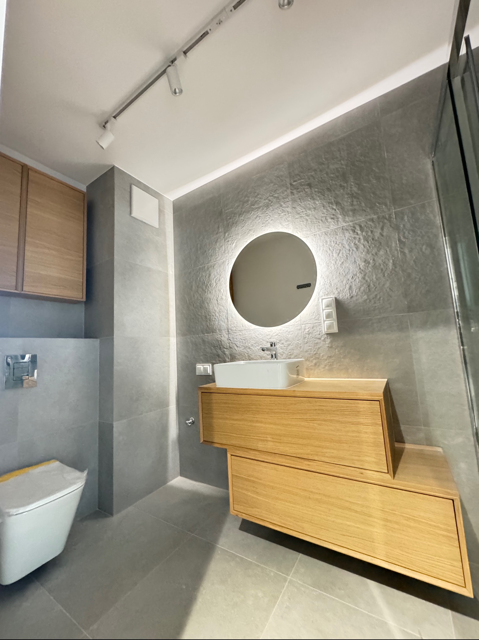 Nowoczesna łazienka w szarościach z drewnianymi akcentami. Uwagę przykuwa podświetlane lustro nad umywalką i oryginalna szafka z szufladami. Minimalistyczny design.
