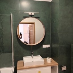 SAD-BUD MATEUSZ SATUŁA - Wykończona łazienka z zielonymi płytkami, okrągłym lustrem i białą szafką pod umywalką. Widoczne akcesoria łazienkowe i odbicie osoby robiącej zdjęcie.