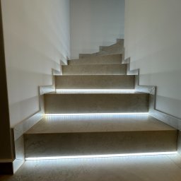 SAD-BUD MATEUSZ SATUŁA - Wykończone schody z kamienia z subtelnym oświetleniem LED, podkreślającym krawędzie stopni. Minimalistyczny design, jasne ściany, nowoczesne wykończenie.