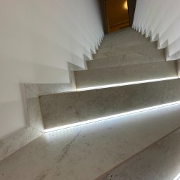 SAD-BUD MATEUSZ SATUŁA - Betonowe schody z subtelnym oświetleniem LED wzdłuż krawędzi stopni, prowadzące w dół. Minimalistyczny design, białe ściany, ciepłe światło w tle.