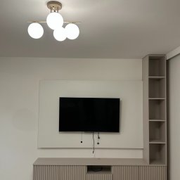 SAD-BUD MATEUSZ SATUŁA - Minimalistyczny salon z beżową szafką RTV z lamelami, wiszącym telewizorem i regałem. Nowoczesna lampa sufitowa z czterema kulami. Jasne wnętrze po remoncie.
