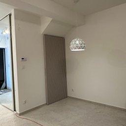 SAD-BUD MATEUSZ SATUŁA - Nowoczesne wykończenie wnętrza: panel ścienny z pionowych listew w odcieniach beżu, designerska lampa wisząca i jasna podłoga z płytek imitujących kamień.