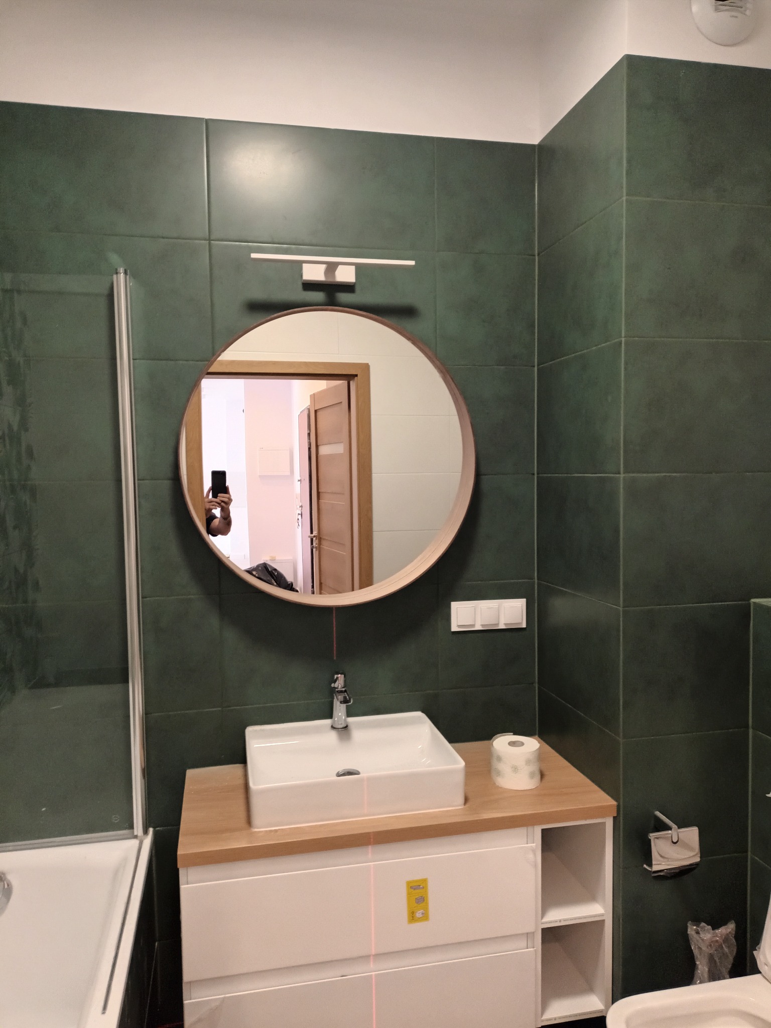 Wykończona łazienka z zielonymi płytkami, okrągłym lustrem i białą szafką pod umywalką. Widoczne akcesoria łazienkowe i odbicie osoby robiącej zdjęcie.