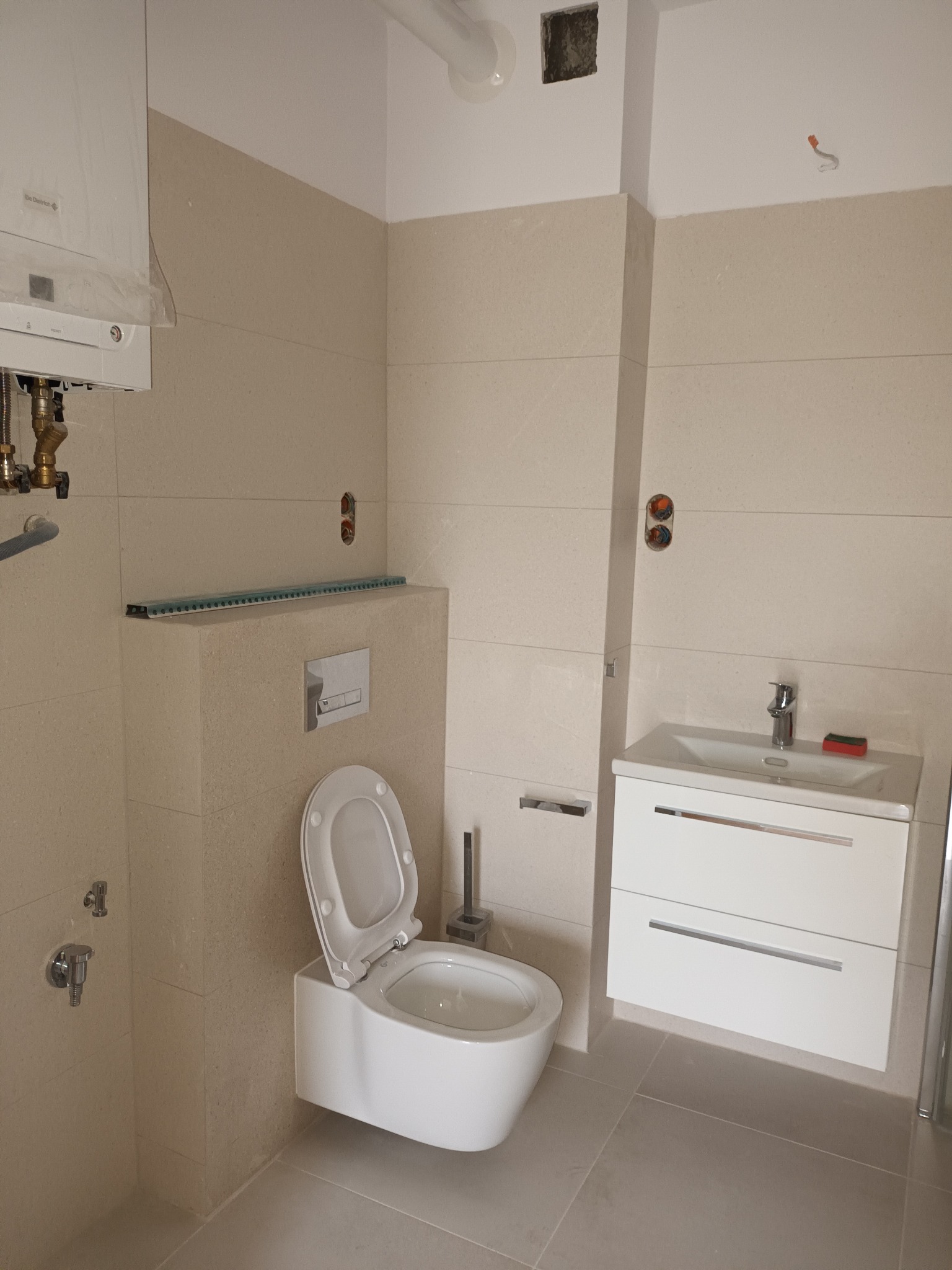 Jasna łazienka w trakcie wykańczania: podwieszana toaleta, umywalka z szafką, beżowe płytki. Widoczne niedokończone elementy instalacji.