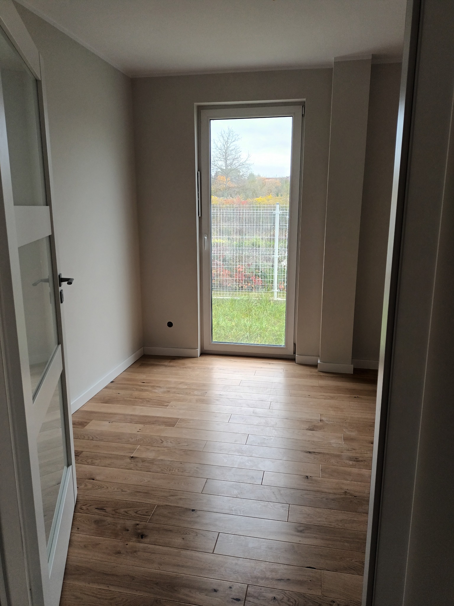 Jasne, puste pomieszczenie z drewnianą podłogą i białymi drzwiami. Widok na zewnątrz przez okno z kratką. Świeżo wykończone wnętrze, gotowe do umeblowania.