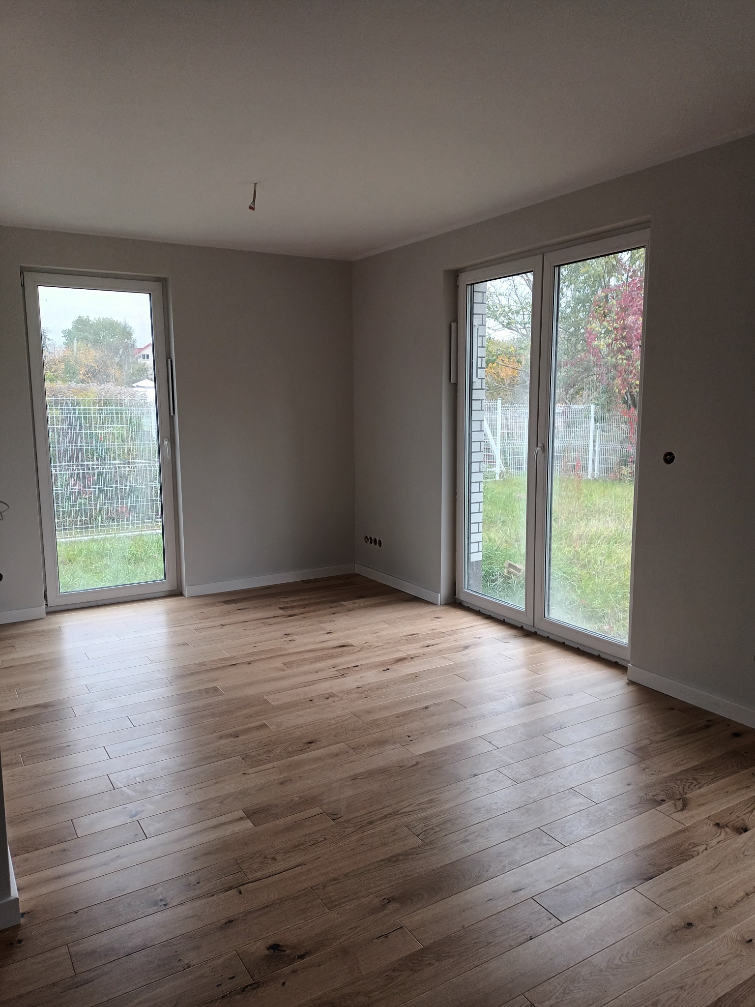 Jasne, drewniane panele podłogowe w pustym pokoju z dwoma oknami wychodzącymi na ogród. Ściany pomalowane na szaro, widoczne gniazdka elektryczne.