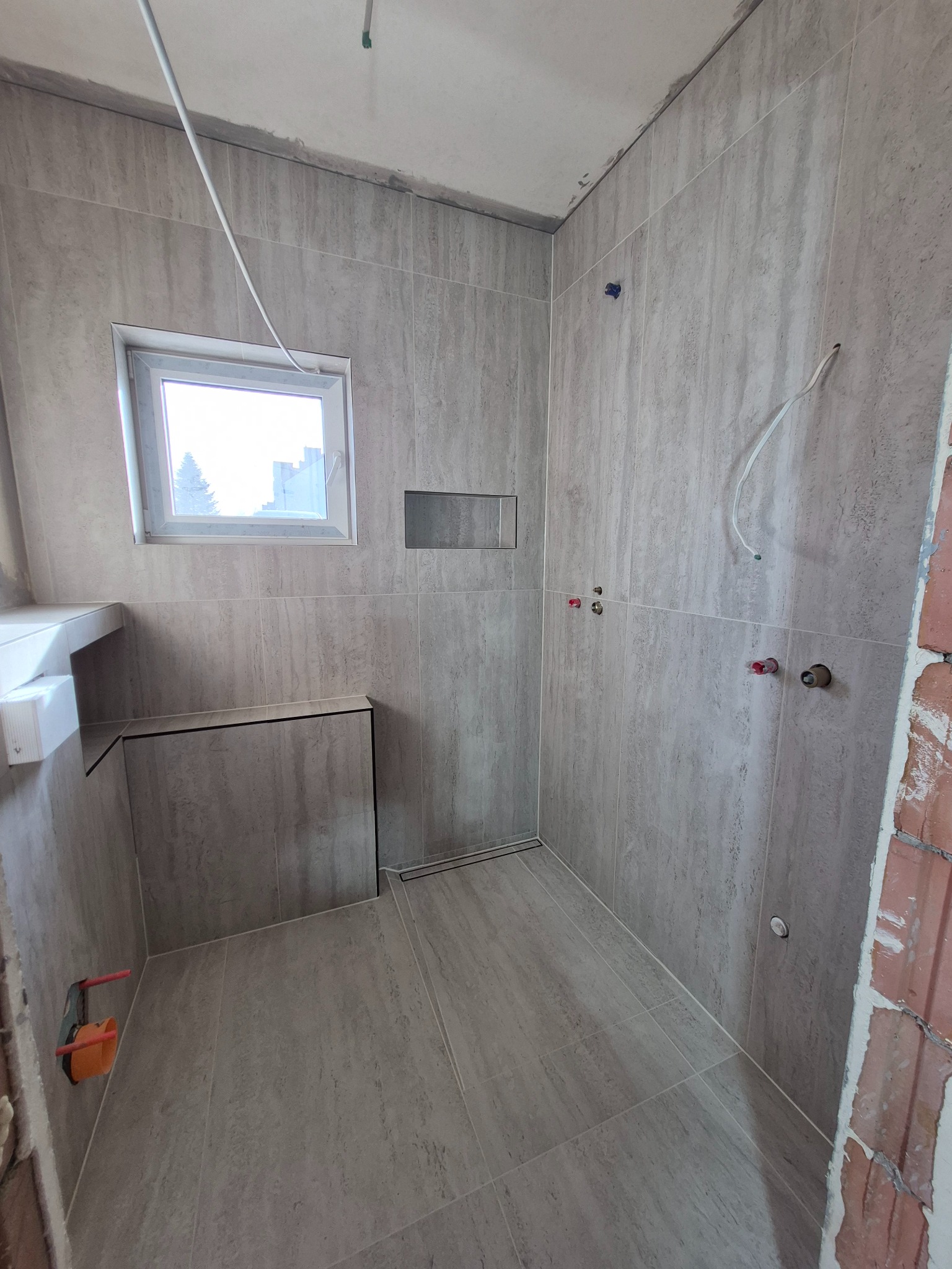 Wykończona łazienka w szarych płytkach imitujących kamień, z oknem i odpływem liniowym. Widoczne wystające kable i rury przed montażem armatury. Surowy mur w rogu.