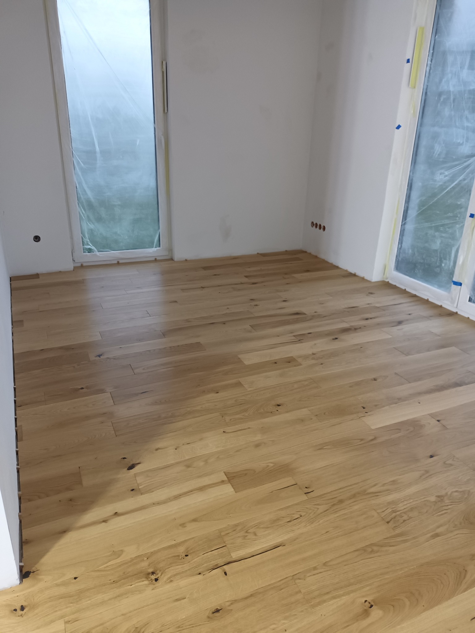 Jasne, drewniane panele podłogowe ułożone w pokoju z oknami zabezpieczonymi folią malarską. Ściany białe, przygotowane do malowania, widoczne gniazdka elektryczne.