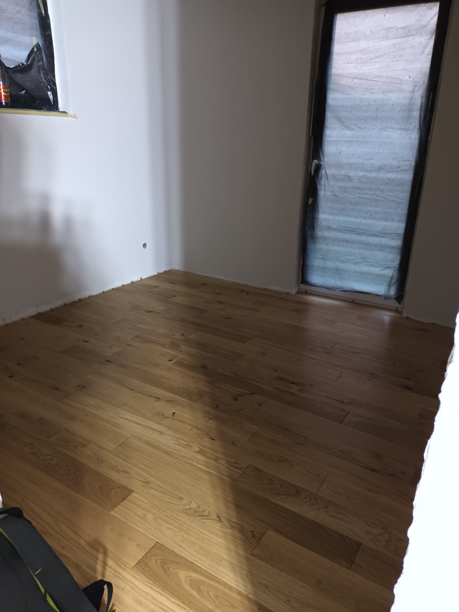 Jasne, drewniane panele podłogowe ułożone w pustym pokoju z oknem zabezpieczonym folią malarską. Widoczne listwy przypodłogowe i fragment torby w rogu.