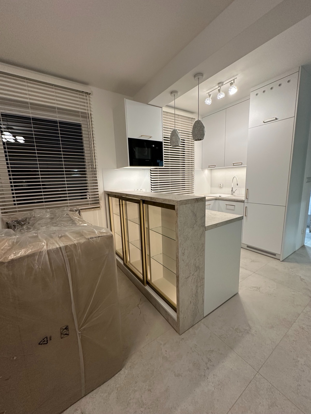 Nowoczesna, biała kuchnia z wyspą i złotymi akcentami. Widoczne żaluzje w oknach i zapakowany karton, sugerujący świeży montaż. Oświetlenie punktowe i marmurowy blat.