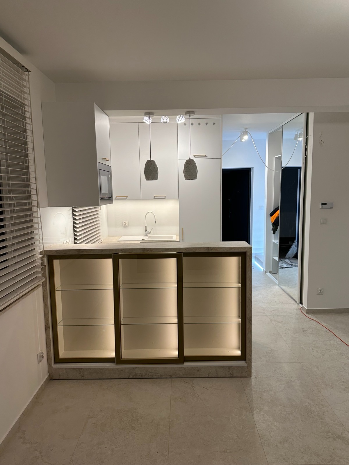 Nowoczesna, jasna kuchnia z białymi szafkami i marmurowym blatem. Podświetlane szklane półki, minimalistyczne lampy wiszące. Widok na lustrzaną szafę w przedpokoju.