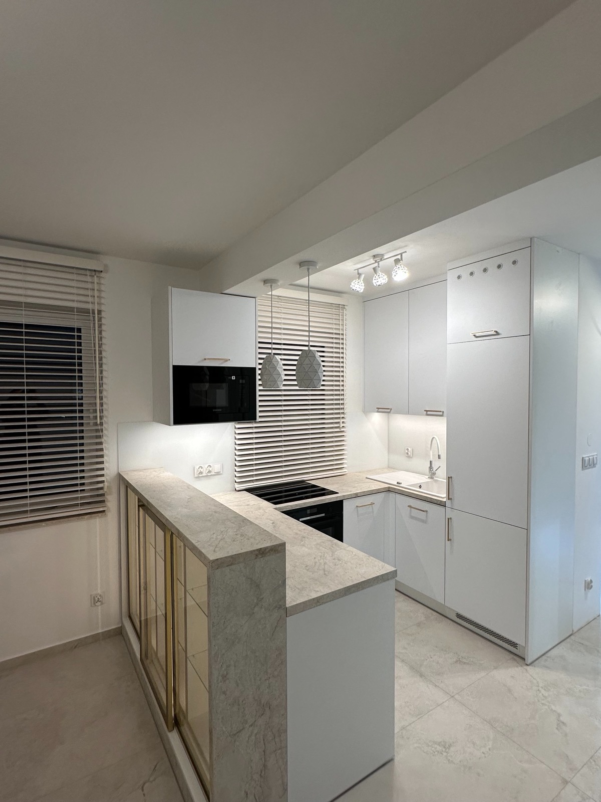 Biała, nowoczesna kuchnia z marmurowym blatem i złotymi akcentami. Widoczne okno z żaluzjami, oświetlenie punktowe i wiszące lampy. Minimalistyczny design.