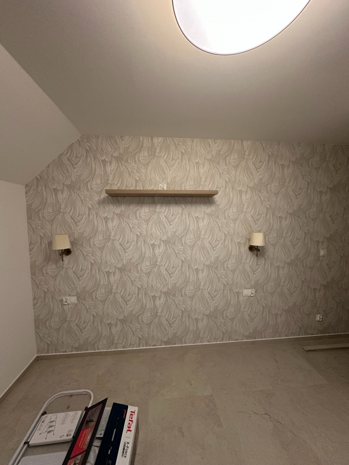 Pokój po tapetowaniu z wzorem liści, z półką i kinkietami. Na podłodze drabina i pudełko. Neutralna kolorystyka, ciepłe oświetlenie sufitowe.