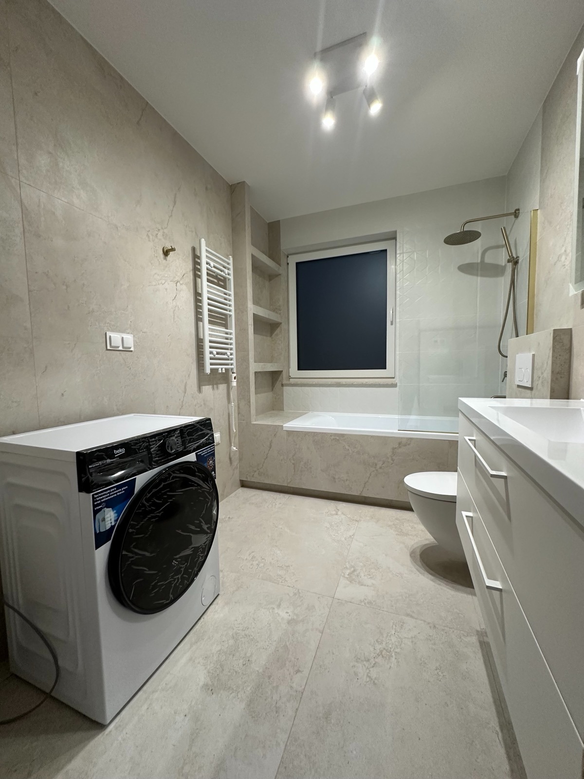 Jasna łazienka z beżowymi płytkami imitującymi kamień, białą pralką Beko z folią ochronną na drzwiczkach, grzejnikiem drabinkowym i zabudowaną wanną.