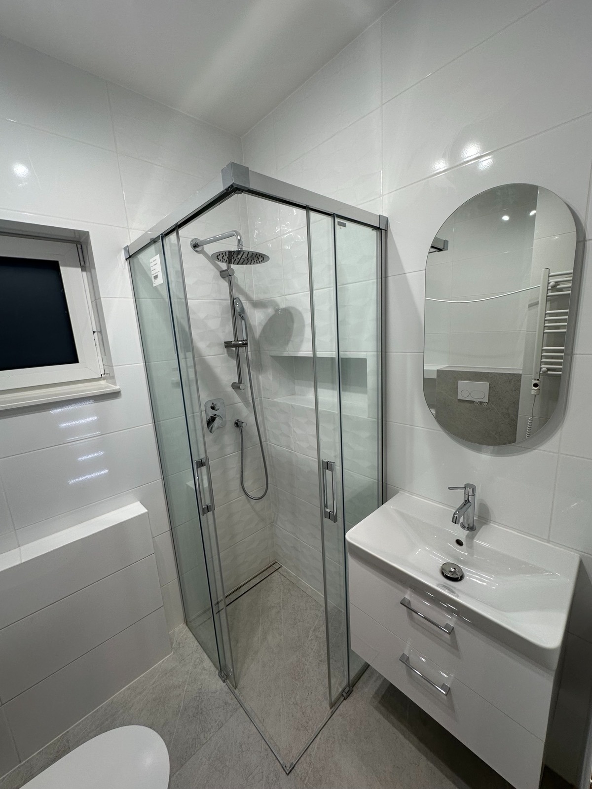 Nowoczesna łazienka po remoncie: kabina prysznicowa z deszczownicą, umywalka z szafką i owalne lustro na tle białych płytek. Minimalistyczny design, jasne wnętrze.