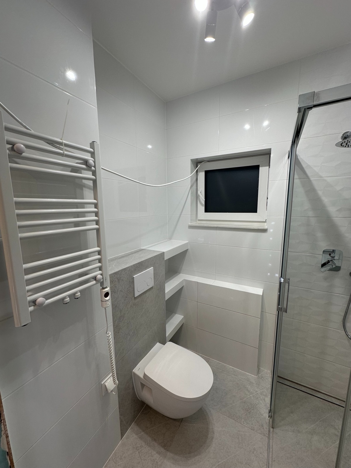 Nowoczesna, jasna łazienka z podwieszaną toaletą, grzejnikiem drabinkowym i kabiną prysznicową. Minimalistyczny design z białymi płytkami i szarymi akcentami.