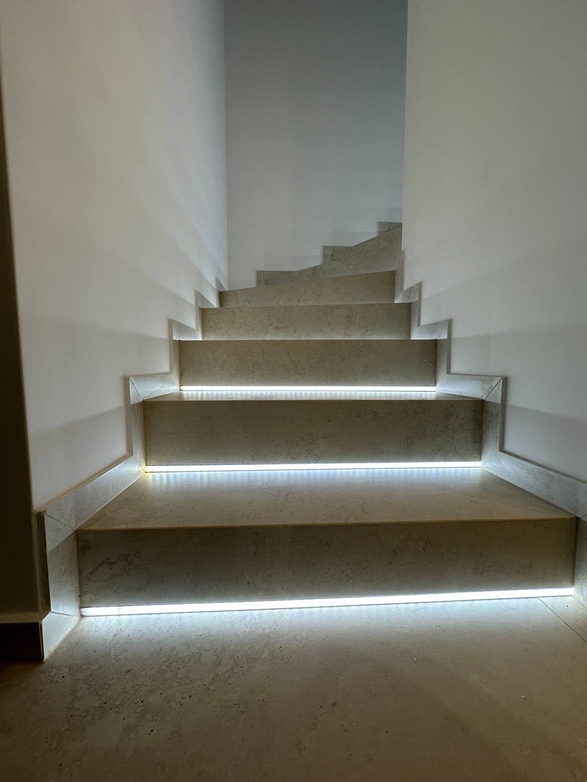 Wykończone schody z kamienia z subtelnym oświetleniem LED, podkreślającym krawędzie stopni. Minimalistyczny design, jasne ściany, nowoczesne wykończenie.