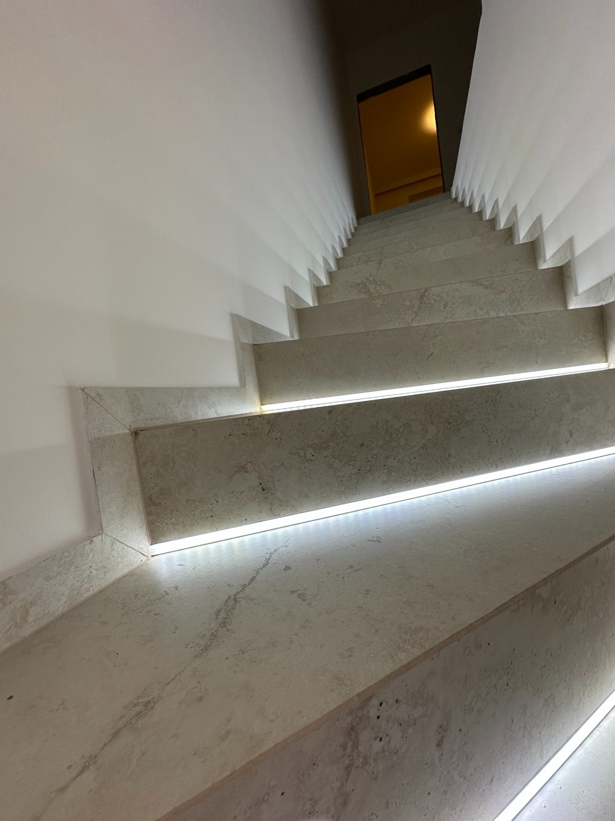 Betonowe schody z subtelnym oświetleniem LED wzdłuż krawędzi stopni, prowadzące w dół. Minimalistyczny design, białe ściany, ciepłe światło w tle.