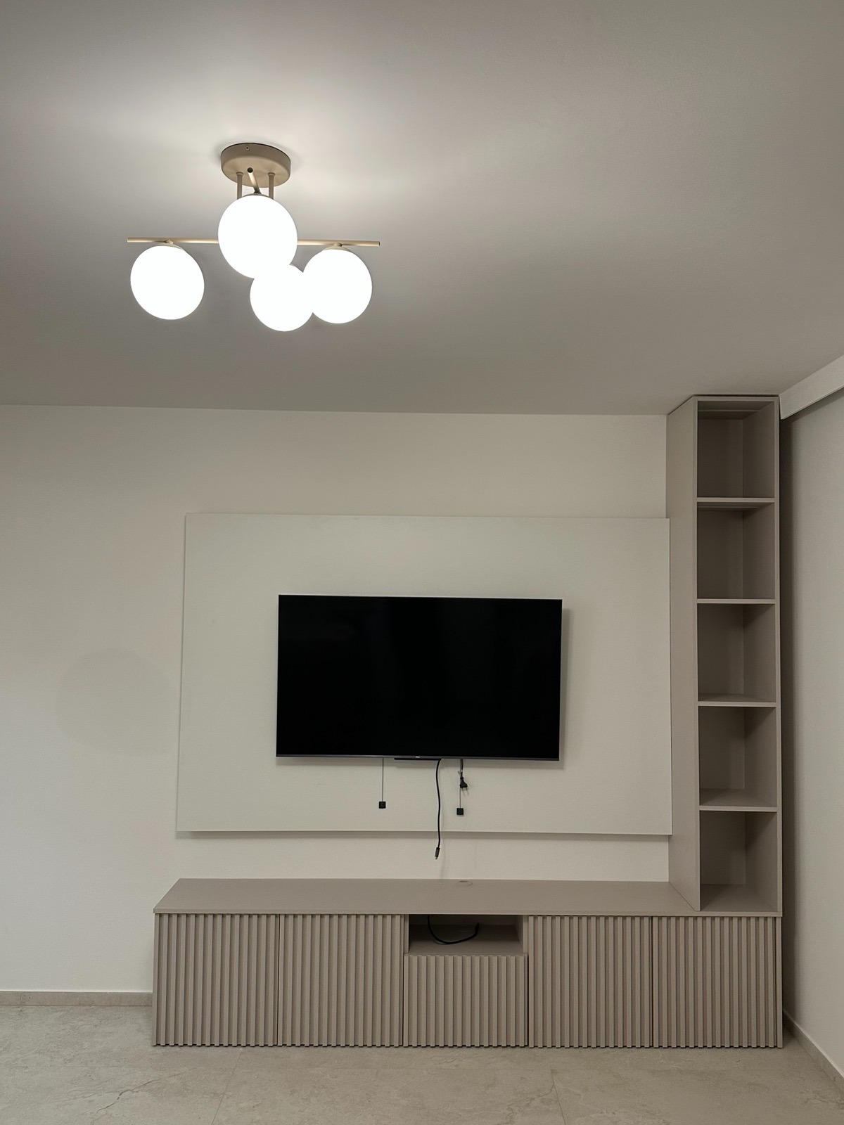 Minimalistyczny salon z beżową szafką RTV z lamelami, wiszącym telewizorem i regałem. Nowoczesna lampa sufitowa z czterema kulami. Jasne wnętrze po remoncie.