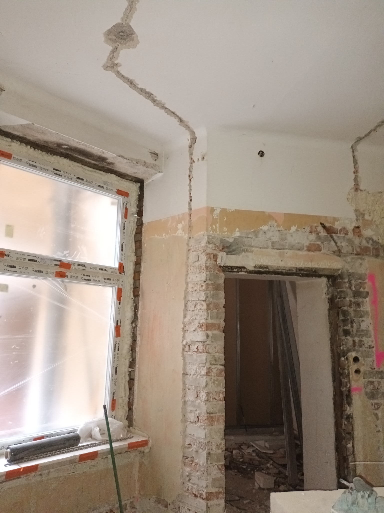 Wnętrze w trakcie demontażu: odsłonięta cegła wokół otworu drzwiowego, okno z folią ochronną, ślady po instalacjach na suficie. Widoczne narzędzia i gruz.