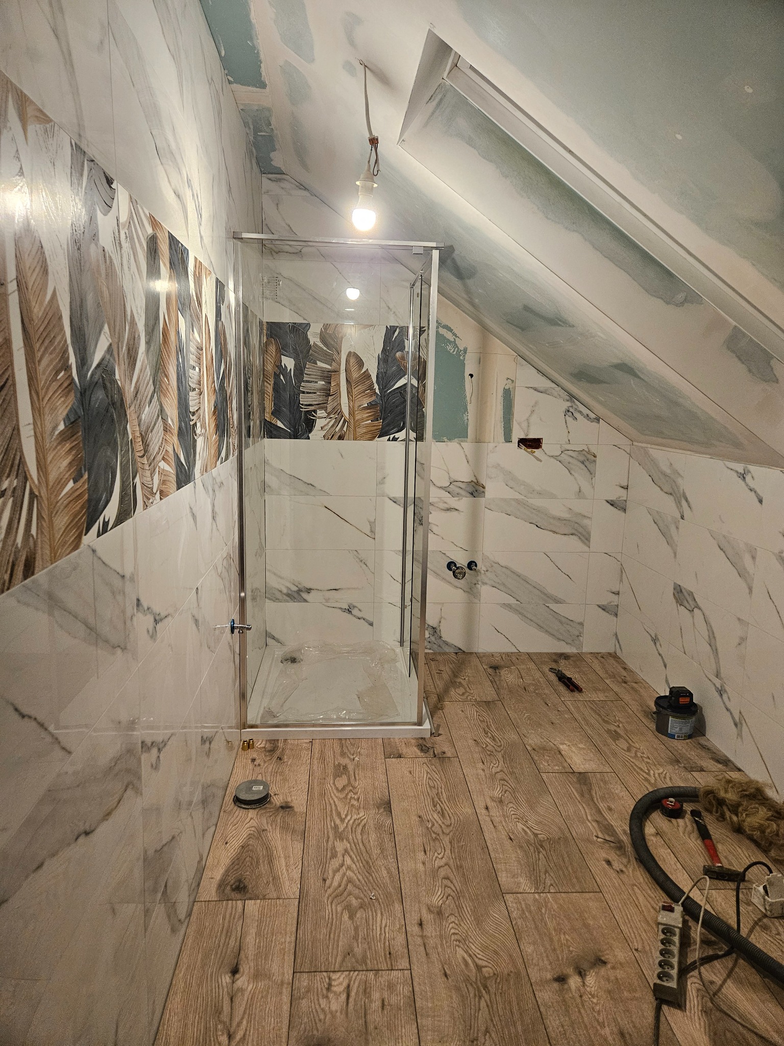 Łazienka na poddaszu w trakcie wykańczania: marmurowe płytki ścienne z motywem liści, drewnopodobna podłoga, kabina prysznicowa ze szkła. Widoczne narzędzia i niedokończone prace.