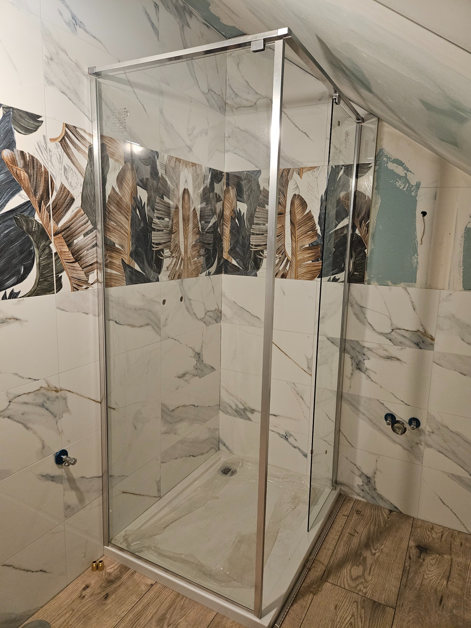 Kabina prysznicowa z chromowanymi profilami, osadzona w łazience z marmurowymi płytkami i motywem liści. Widoczne elementy instalacji wodnej i drewnopodobna podłoga.