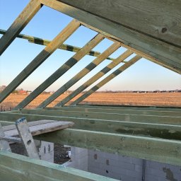 NFO-DACH Filip Wróblewski - Więźba dachowa z zielonego drewna na tle błękitnego nieba i pola. Widoczne białe bloczki fundamentowe i drabina. Kąt ujęcia od dołu, podkreślający konstrukcję dachu.