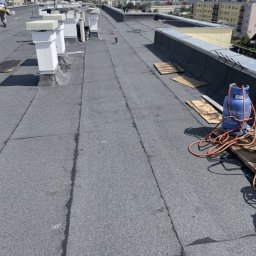 NFO-DACH Filip Wróblewski - Długi, płaski dach pokryty papą, z kominami i butlą gazową. Widoczne miejskie budynki w tle. Prace dekarskie w toku, z użyciem palnika.