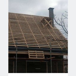 NFO-DACH Filip Wróblewski - W trakcie budowy dachu: widok na drewnianą konstrukcję więźby z łatami, komin z drewnianym rusztowaniem, rusztowania i fragment drzewa na tle pochmurnego nieba.