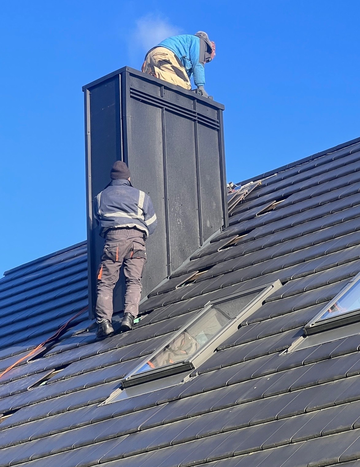 Praca dekarzy na dachu z oknami połaciowymi i kominem w Stargardzie. Dwóch mężczyzn w odzieży roboczej wykonuje prace na wysokości, na tle błękitnego nieba.