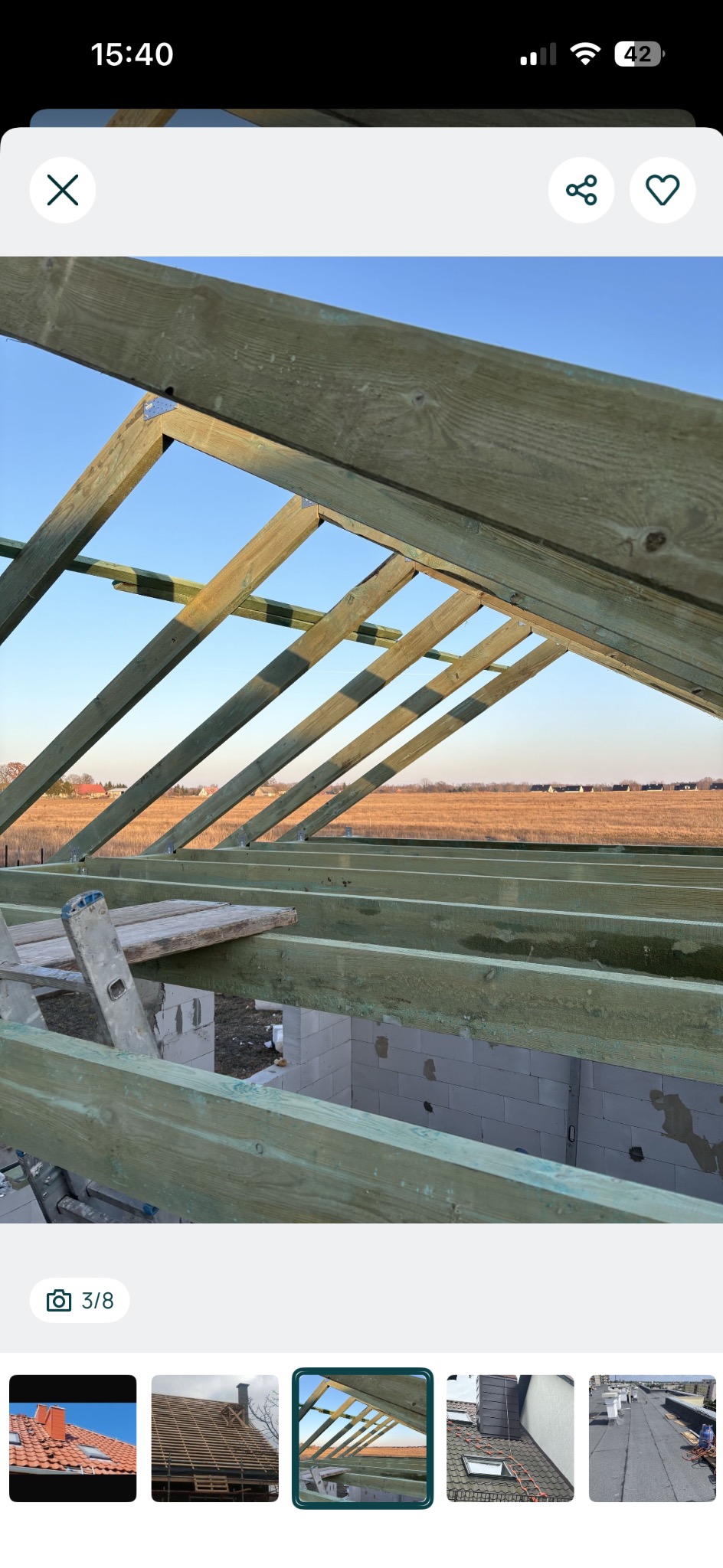 Więźba dachowa z zielonego drewna na tle błękitnego nieba i pola. Widoczne białe bloczki fundamentowe i drabina. Kąt ujęcia od dołu, podkreślający konstrukcję dachu.