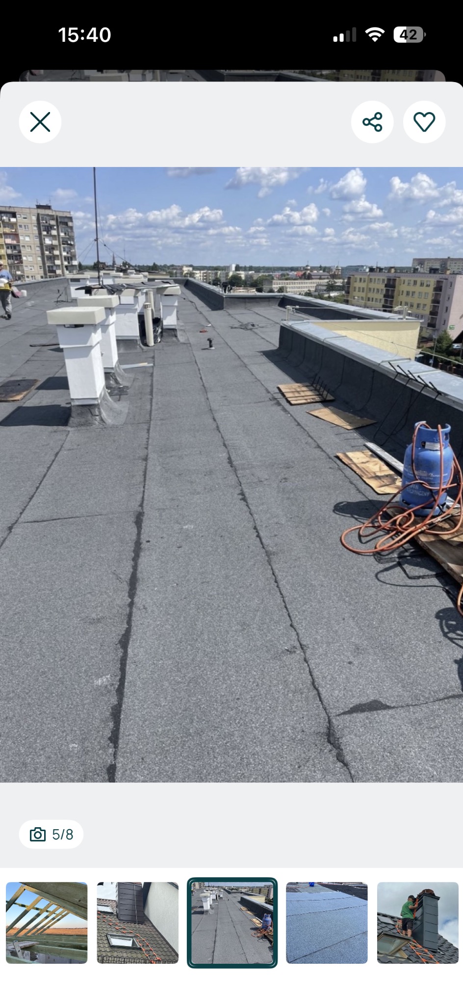 Długi, płaski dach pokryty papą, z kominami i butlą gazową. Widoczne miejskie budynki w tle. Prace dekarskie w toku, z użyciem palnika.