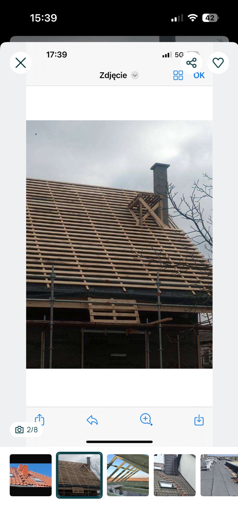 W trakcie budowy dachu: widok na drewnianą konstrukcję więźby z łatami, komin z drewnianym rusztowaniem, rusztowania i fragment drzewa na tle pochmurnego nieba.