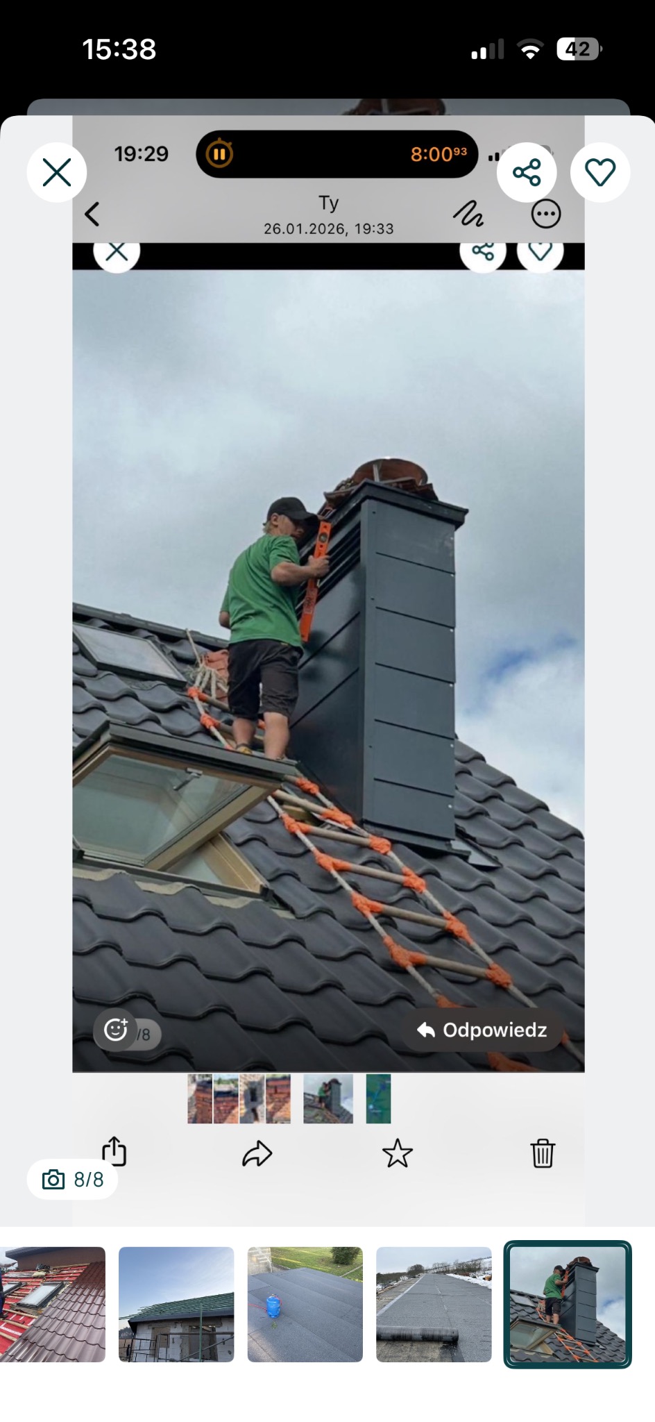 Pracownik na dachu z drabinką kominiarską, montuje obróbkę komina z blachy w kolorze grafitowym. Dachówka ceramiczna w kolorze antracytowym. Widoczne okno dachowe.