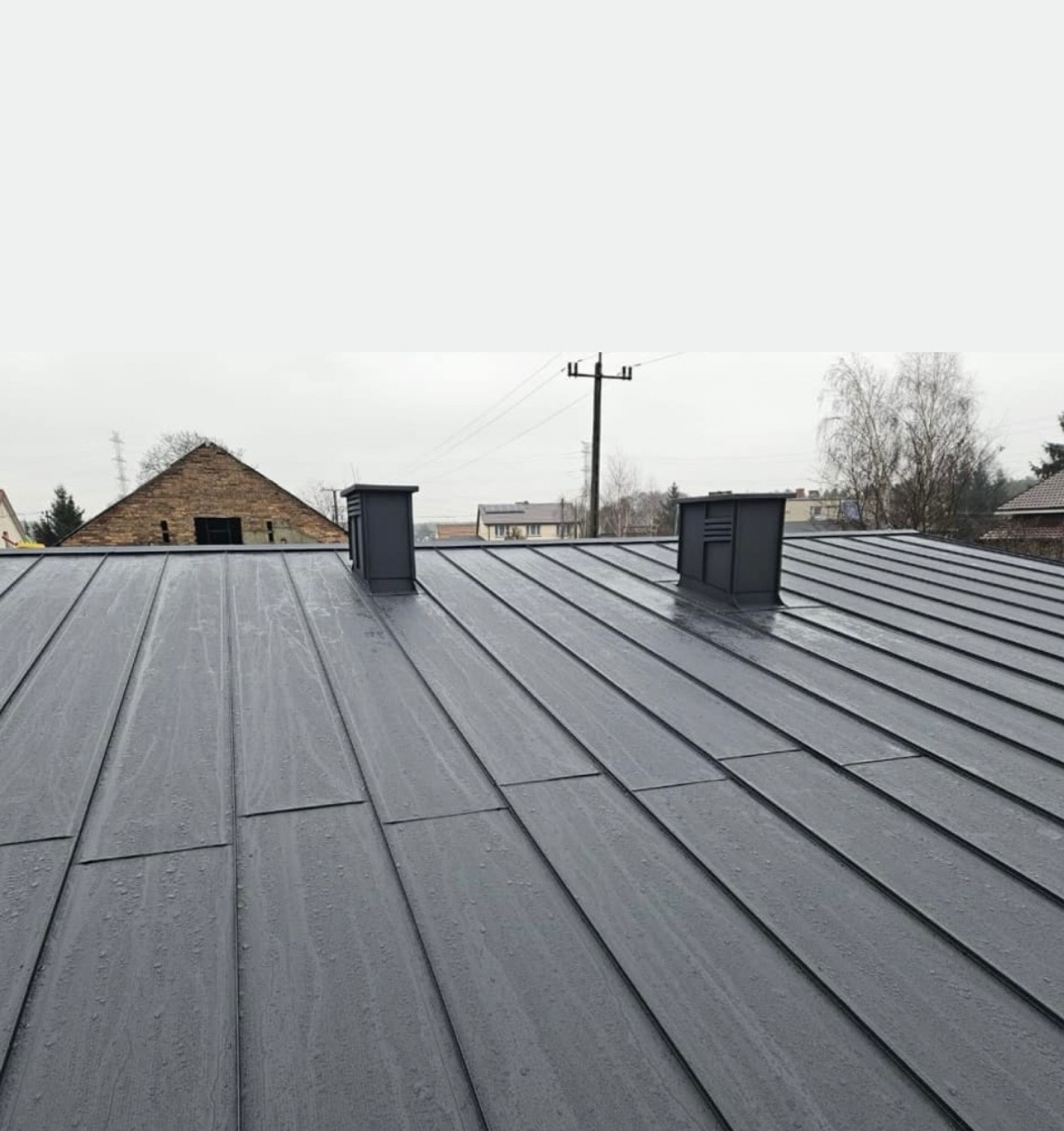Szary, metalowy dach panelowy z dwoma kominami wentylacyjnymi na tle pochmurnego nieba i zabudowań. Widoczne ślady wilgoci na powierzchni dachu.