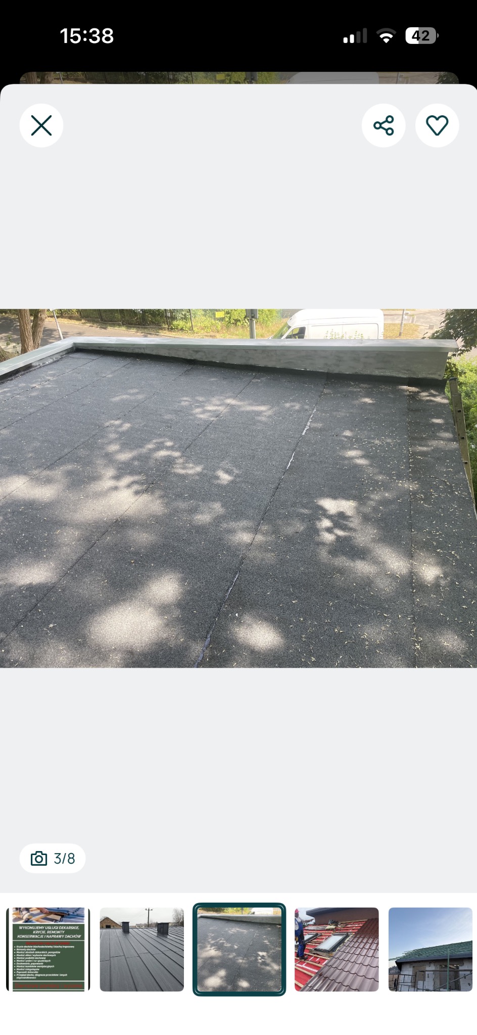 Płaski dach pokryty papą, widoczne łączenia i obróbki blacharskie. Na powierzchni dachu leżą drobne zanieczyszczenia. W tle biały samochód dostawczy.